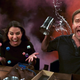 Cast members of Critical Role at the role-playing table, with dice in the air and liquid spilling from a tankard as one player cheers.